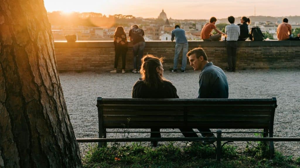 A couple sits apart on a park bench at sunset, with other people in the background.