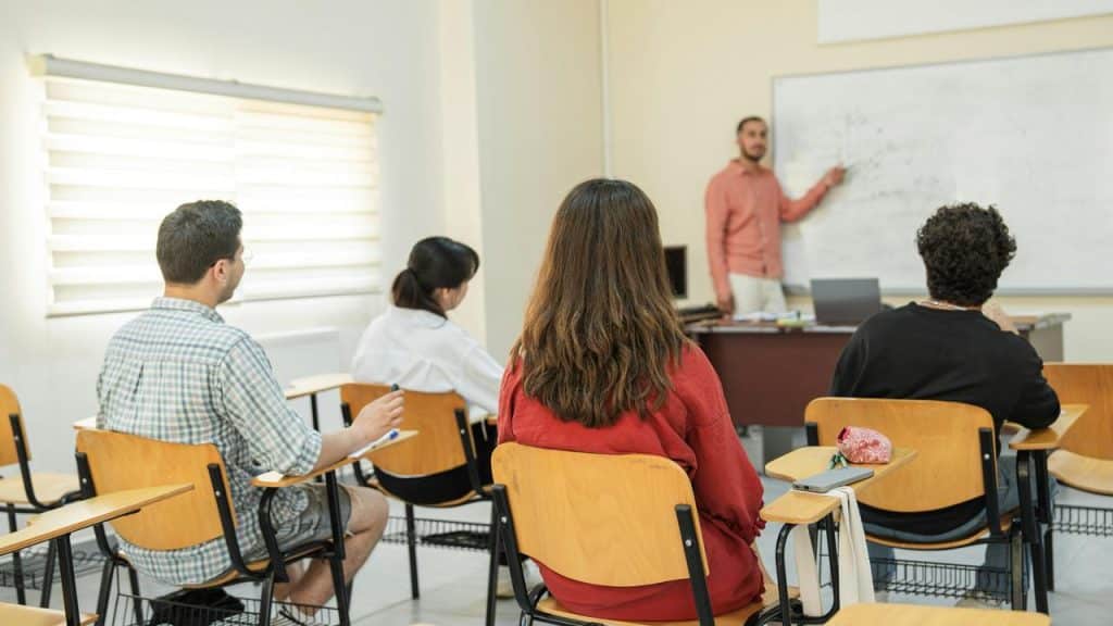 A classroom of students watching a teacher write on a whiteboard.