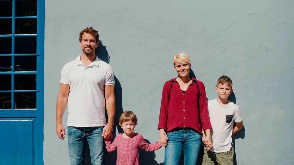 A man, a woman, and two children standing together, holding hands in front of a blue wall.