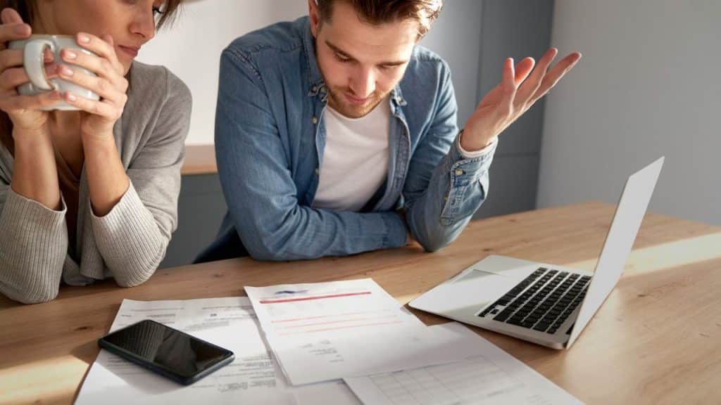 A frustrated couple sitting at a table with bills and a laptop, with the man gesturing in exasperation.
