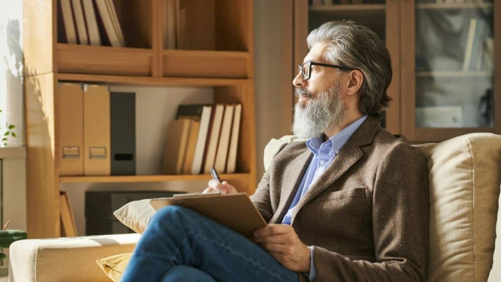 A bearded man in glasses sits on a couch, writing on a clipboard and looking off into the distance.