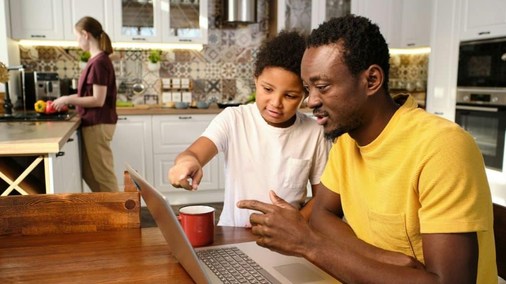 A father and son looking at a laptop at a kitchen table, with a woman in the background.
