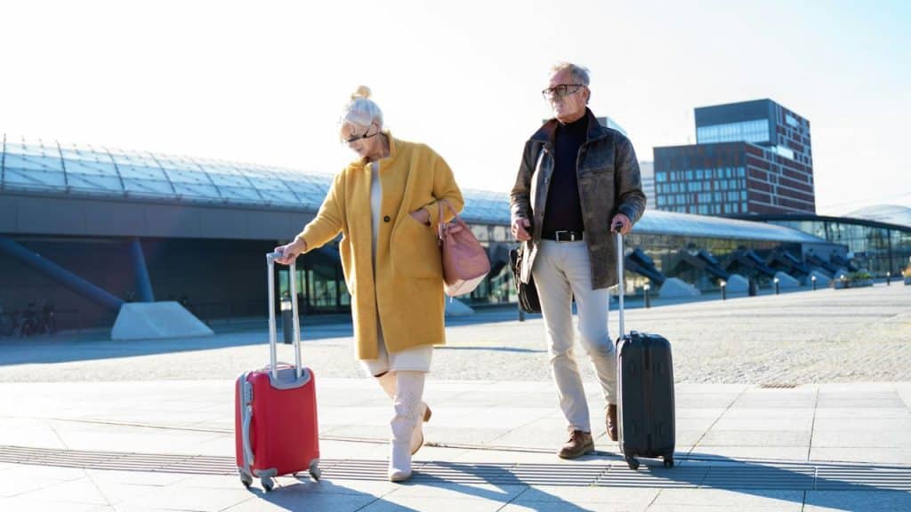 An older couple walking away from a building, pulling their luggage behind them.