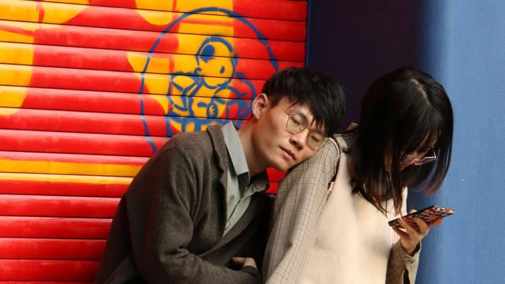 A couple standing together against a wall, with the man leaning his head on the woman's shoulder as she looks at her phone.