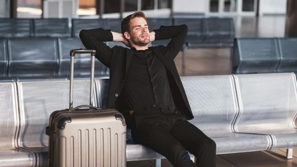 A man wearing a suit leans back with his hands behind his head in an airport waiting area.