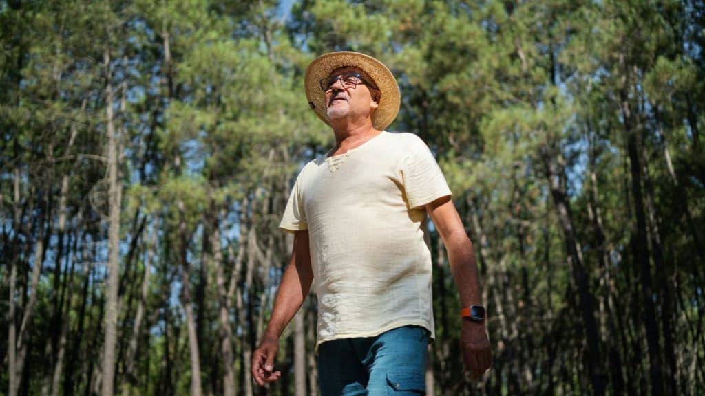 A content man wearing a straw hat and glasses, walking in a forest and looking up at the sky.