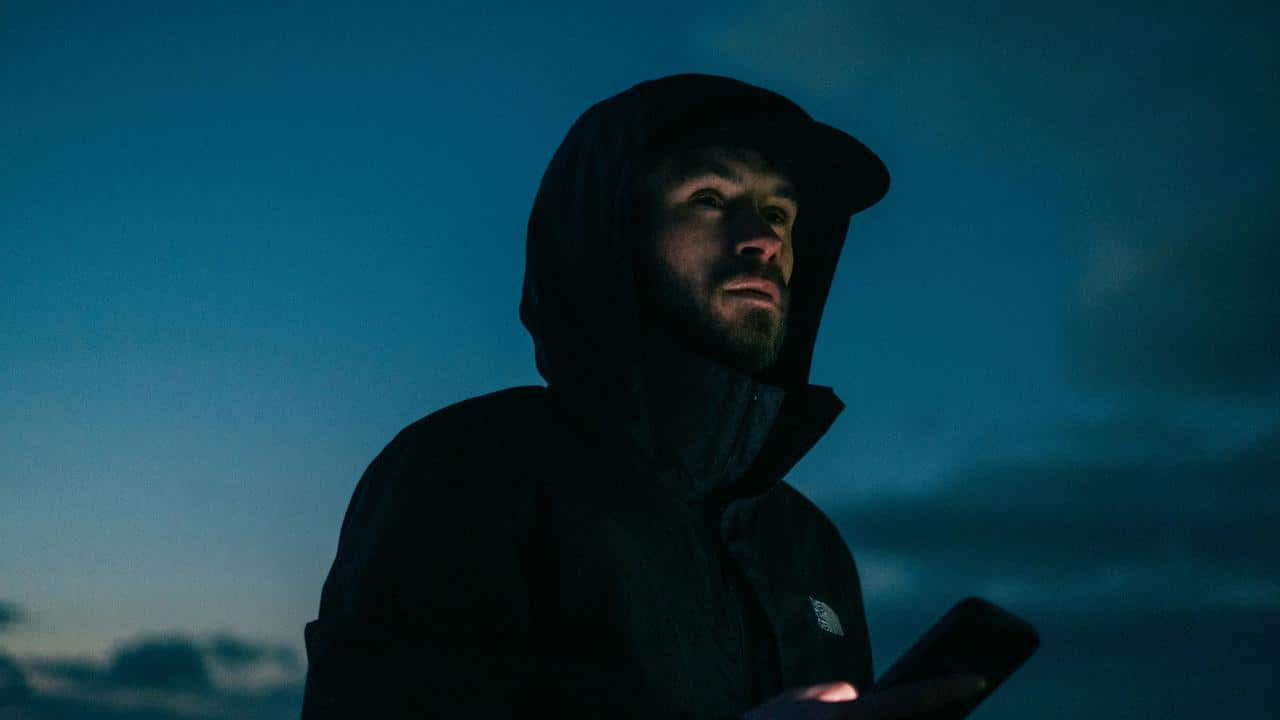 A man wearing a dark hooded jacket holding a phone at dusk.