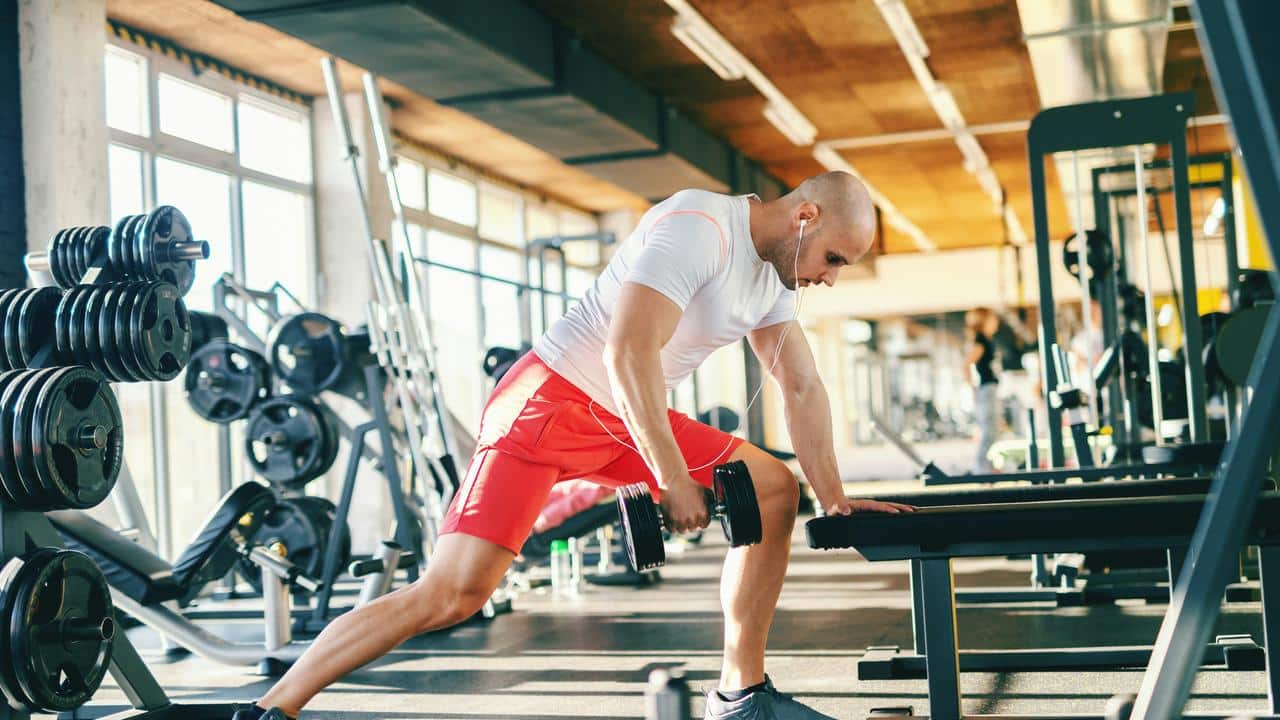 A man in red shorts and a white shirt lifting a dumbbell in a gym.