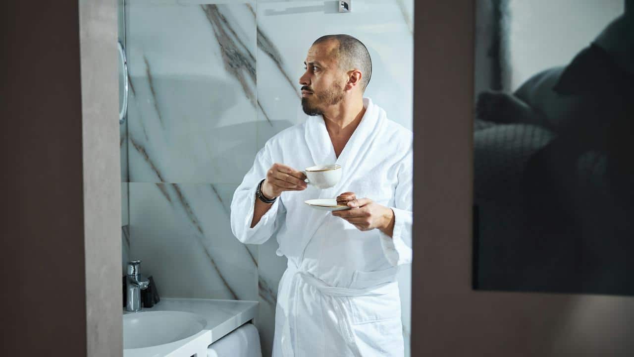 A man in a white bathrobe holding a cup and saucer in a bathroom.