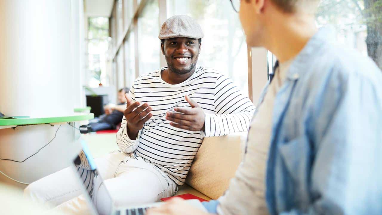 A man in a striped shirt and cap smiling while talking to another person indoors.