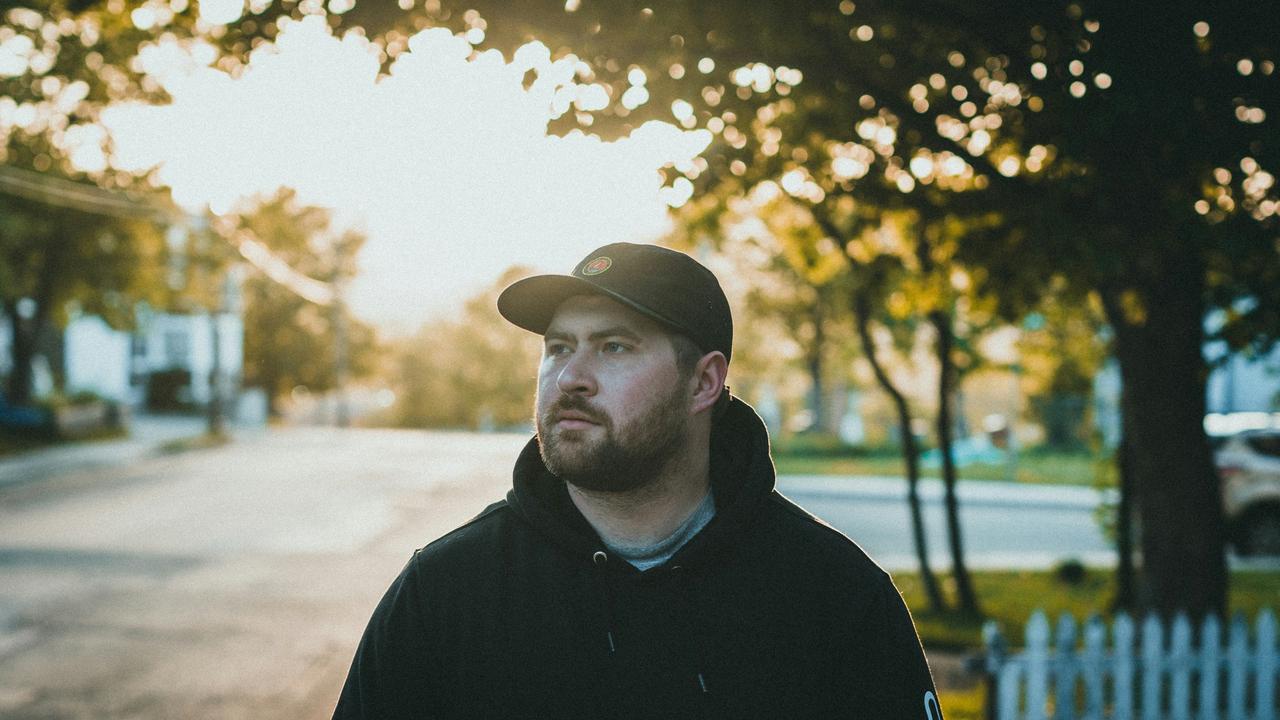 A man in a black hoodie and cap standing on a street at sunset.