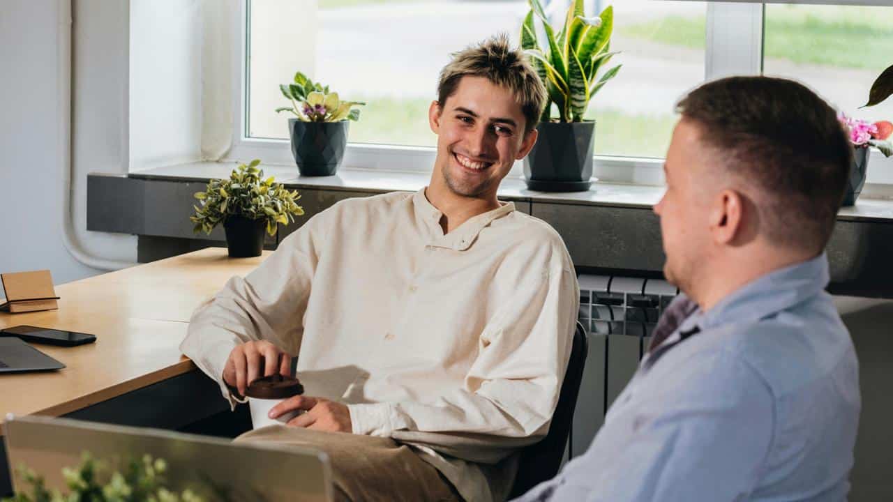 A pair of men smiling and talking at an office desk.