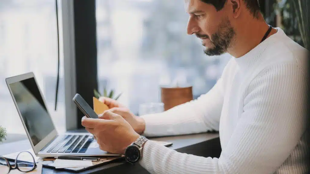 A man using a smartphone while seated at a desk with a laptop.