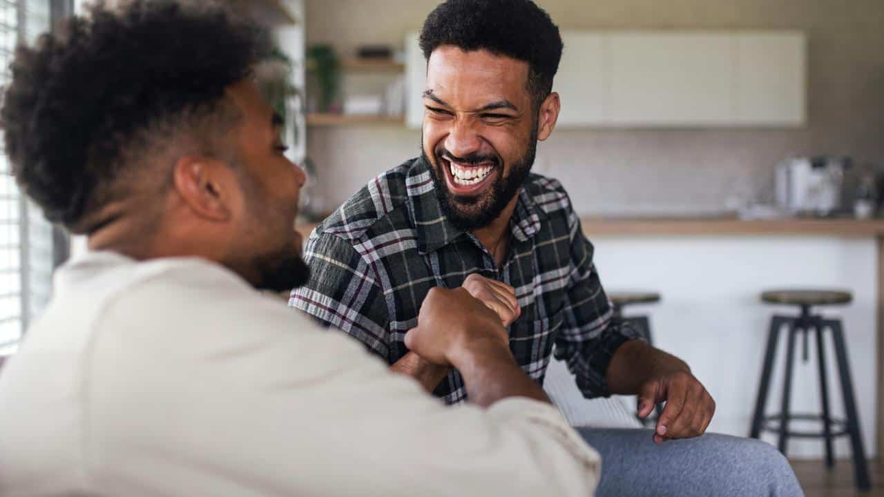 A pair of men laughing and shaking hands in a kitchen.