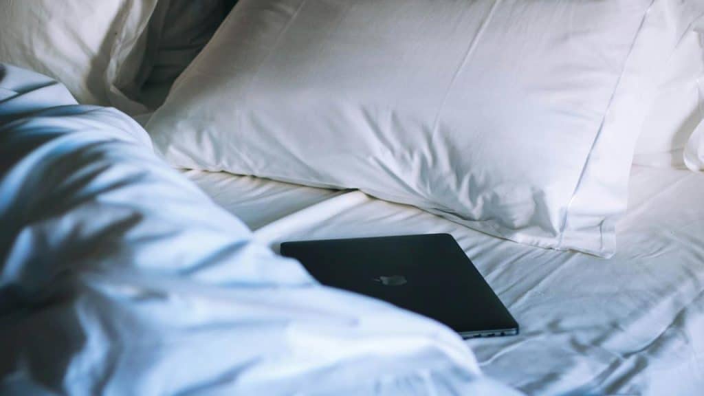 A closed laptop resting on rumpled white bed sheets.