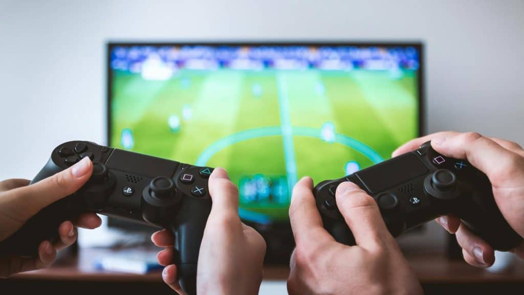 A pair of hands holding PlayStation controllers in front of a TV showing a soccer video game.