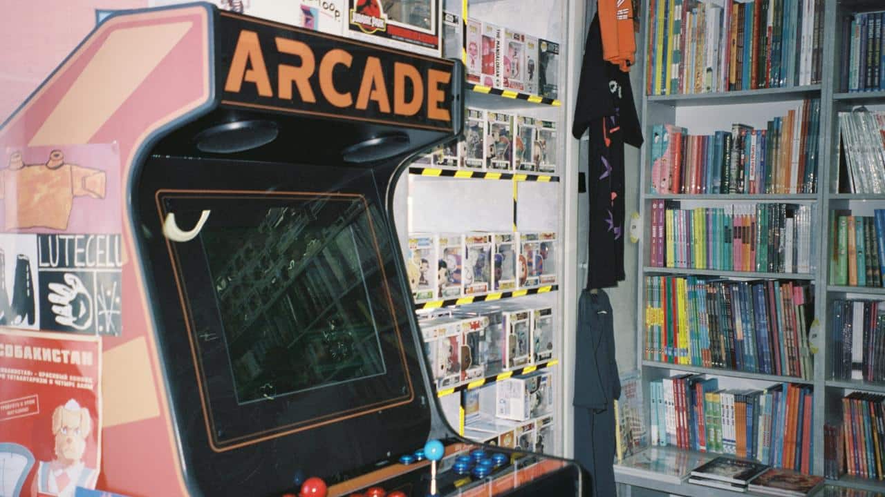 An arcade machine beside shelves of comics, collectibles, and tees.