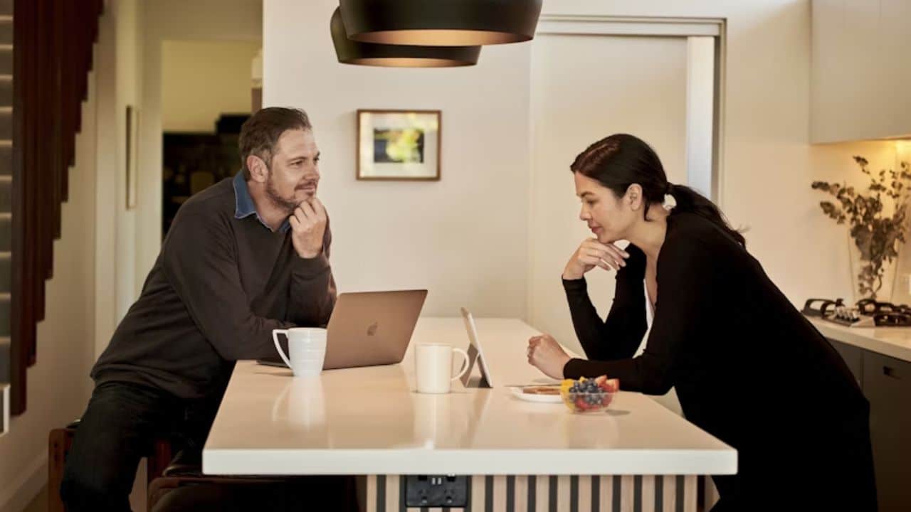 A couple sitting at home with a laptop and coffee mugs, smiling while checking their finances together.
