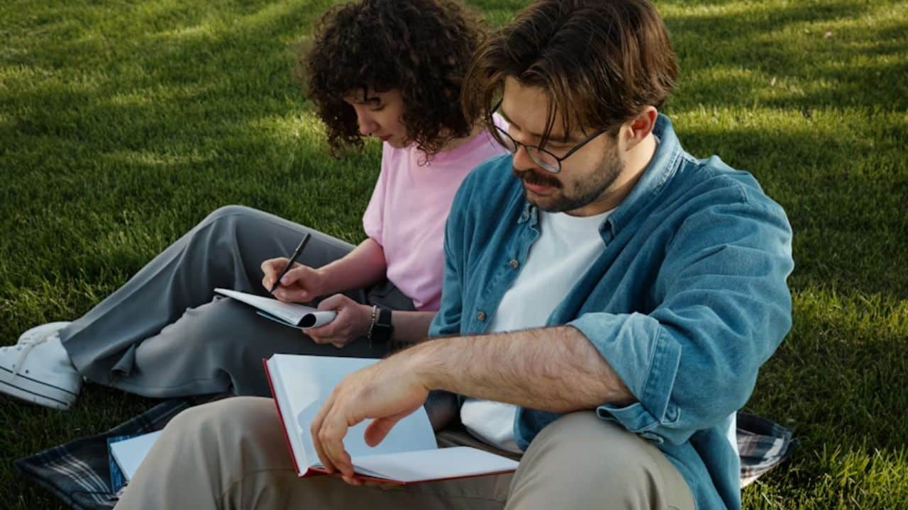 Man reading a book while his partner sits beside him taking notes.