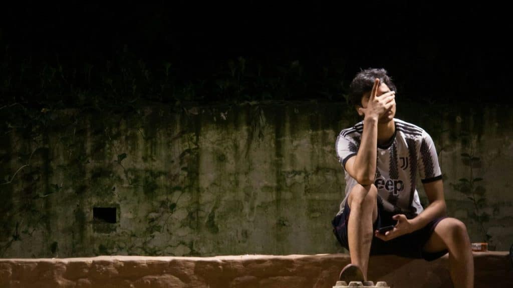 A worried man is covering his face with his hand while sitting outside at night.