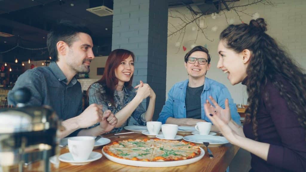 A group of friends is sitting at a table with a pizza and coffee cups, laughing and talking.