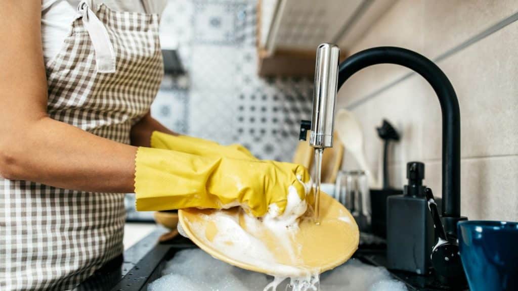 A person wearing a checkered apron and yellow gloves is washing a plate in a sink.