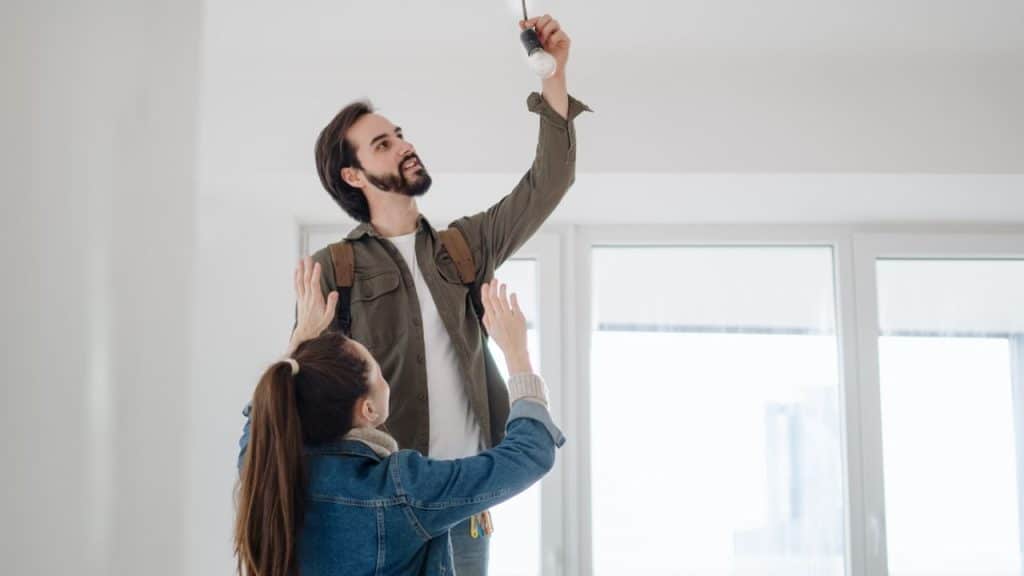 A couple is in a room with a man on a ladder changing a light bulb, and a woman is holding a screwdriver and gesturing.