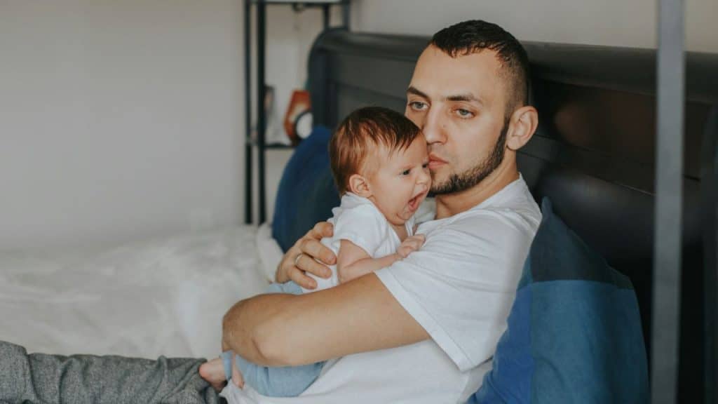 A concerned father with a beard is holding a crying baby in his arms.
