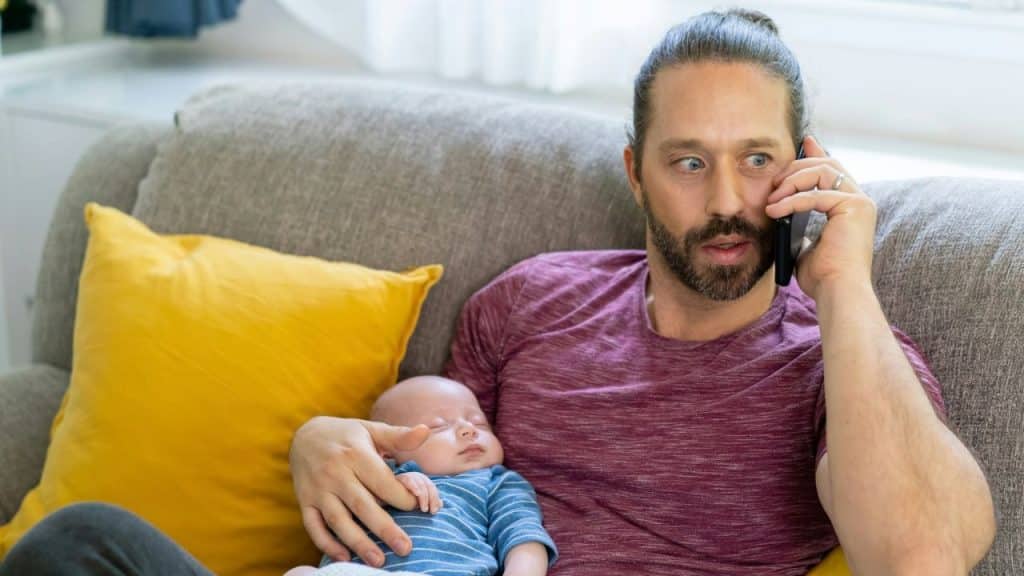A surprised father is holding a sleeping baby in his arms while talking on the phone.