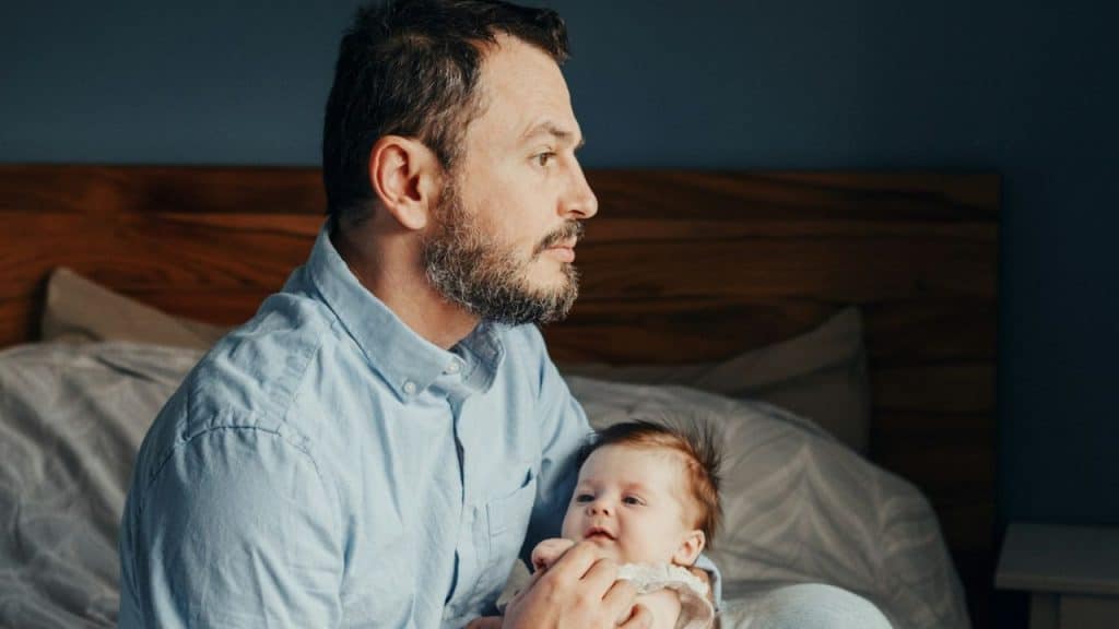 A worried father is holding his baby, looking away with a concerned expression.