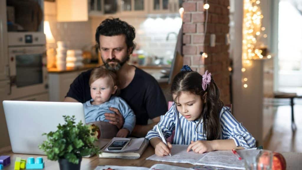 A busy father with a baby on his lap is working on a laptop while his young daughter is sitting next to him and doing homework.