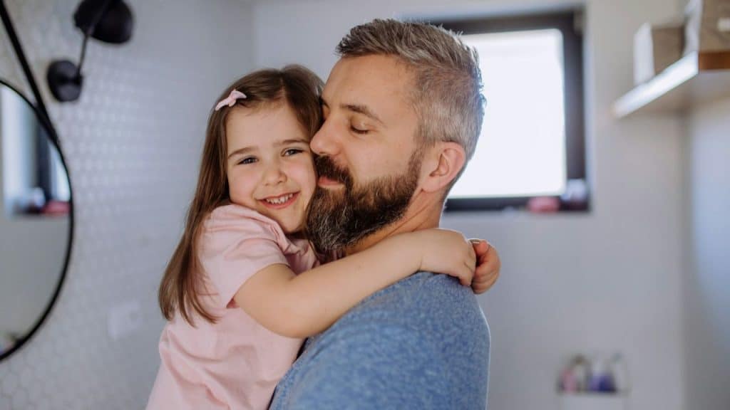 A smiling father holds his joyful daughter, hugging her in a bathroom.