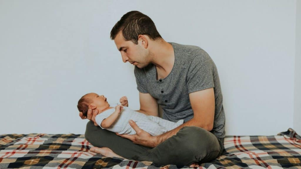 A man sits cross-legged, holding and looking at a newborn baby.