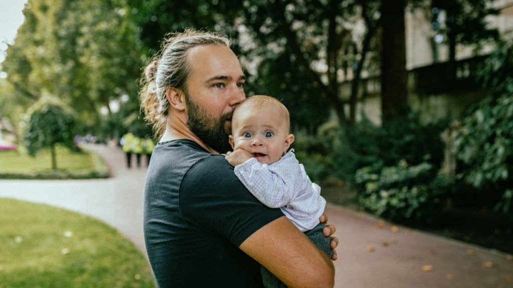 A man with long hair in a bun holds a wide-eyed baby outdoors.