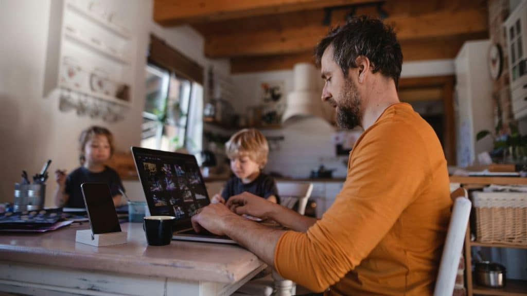 A man with a beard works on a laptop at a kitchen table. His two children are in the background.