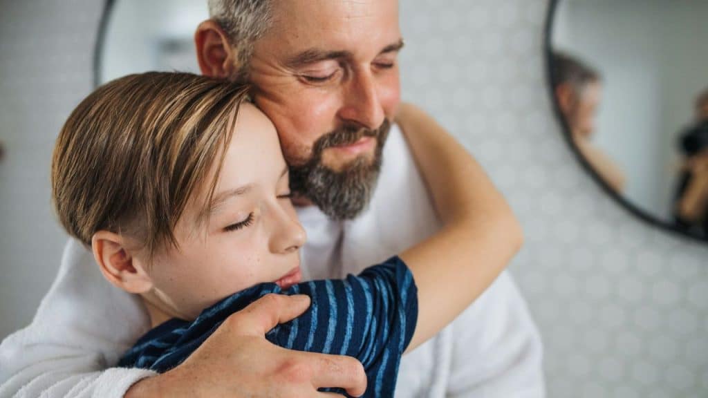 A smiling father hugs his son with his eyes closed in a bathroom.