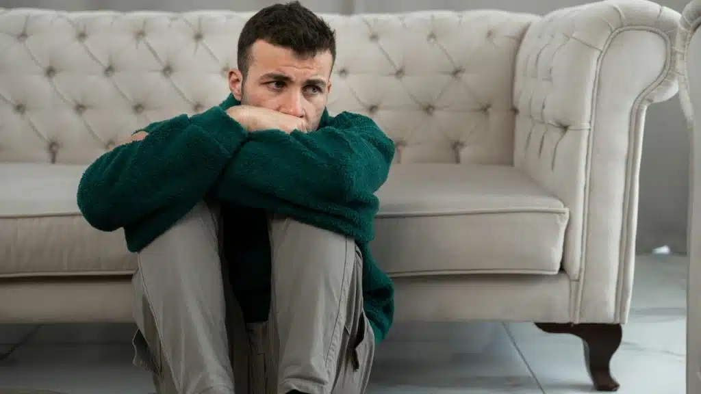 A man sits on the floor, hugging his knees with his arms and looking to the side.