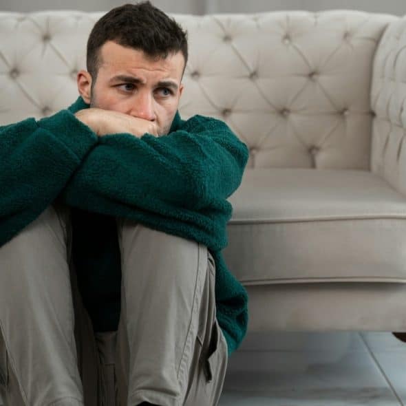A man sits on the floor, hugging his knees with his arms and looking to the side.