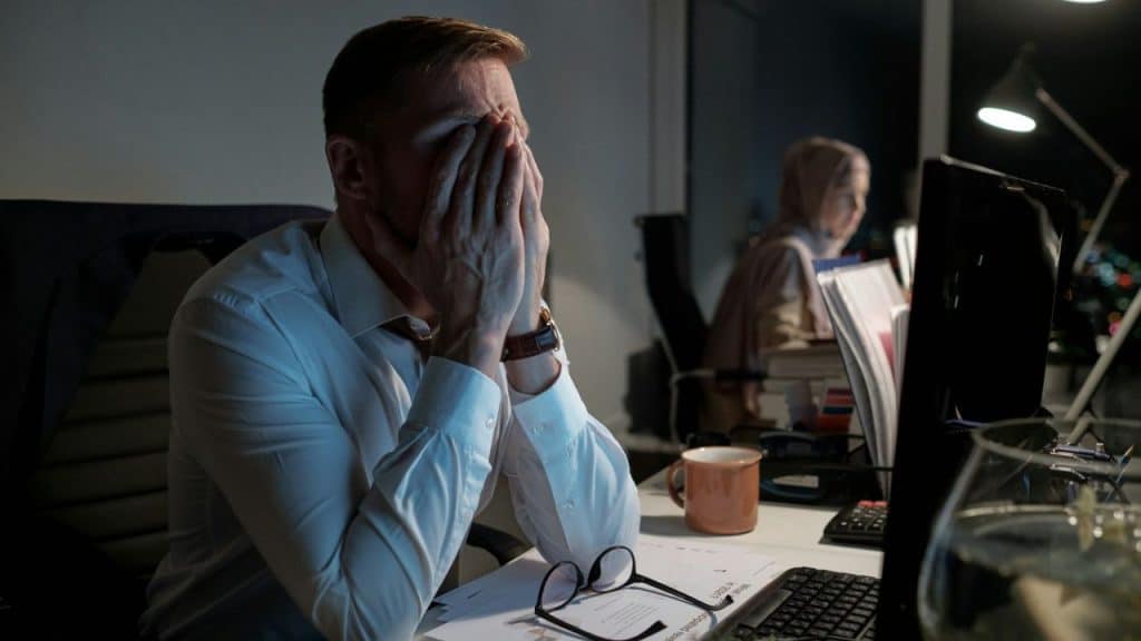 A man in an office covers his face with his hands while looking at a computer screen.