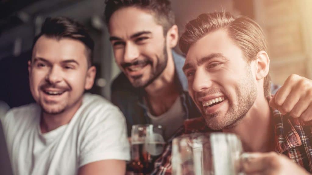 Three men smile while looking at a smartphone and drinking beer.