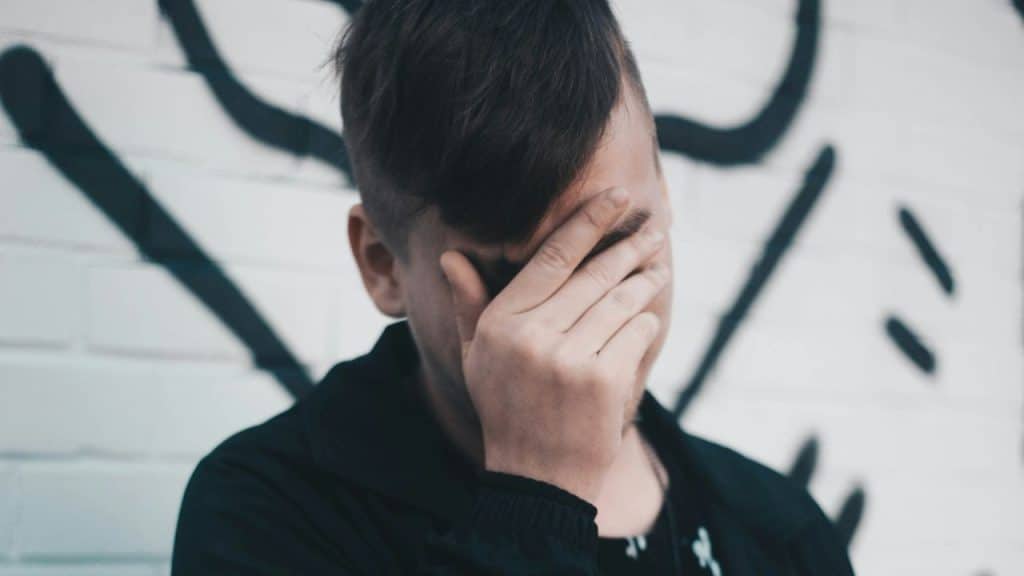 A man covers his face with his hand while standing near a wall with graffiti.