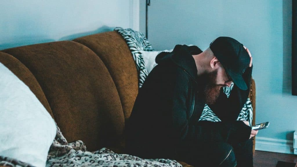 A man wearing a baseball cap and a beard sits on a couch, holding his head while looking at his smartphone.