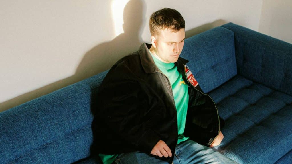 A man in a black jacket and a green shirt sits on a blue couch.
