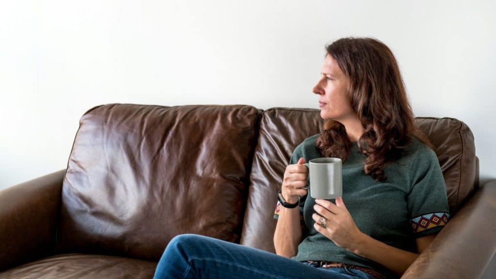 A woman sits on a leather couch, holding a mug and looking to her right.