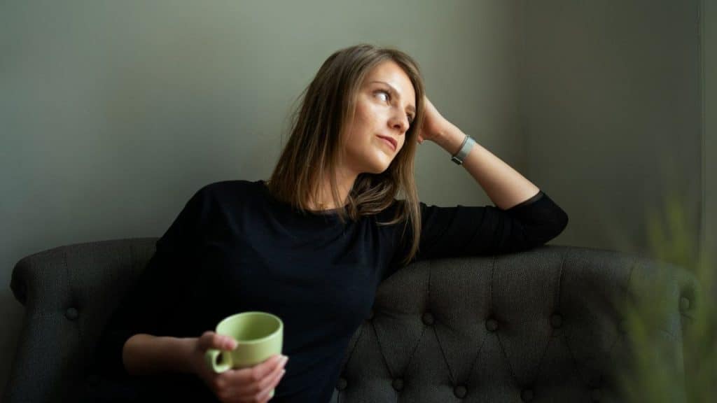A woman sits on a couch, holding a mug and looking away from the camera.