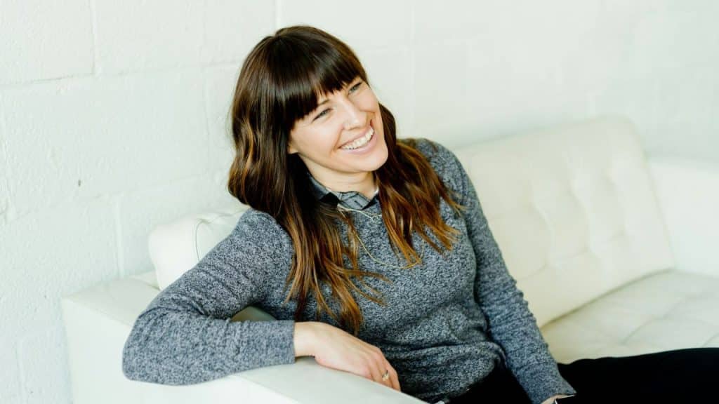 A woman with bangs and brown hair sits on a white couch and smiles.