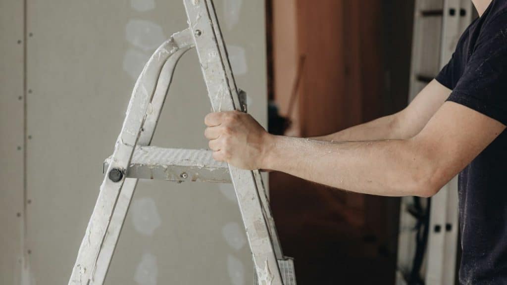 A person's hands grip a ladder with paint or drywall dust on it.