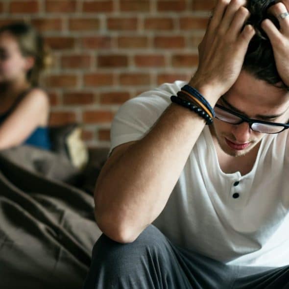 A man sits on a bed with his head in his hands, while a woman looks at her phone in the background.