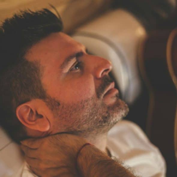 A handsome man with a beard and dark hair is lying on a white couch, looking pensive.