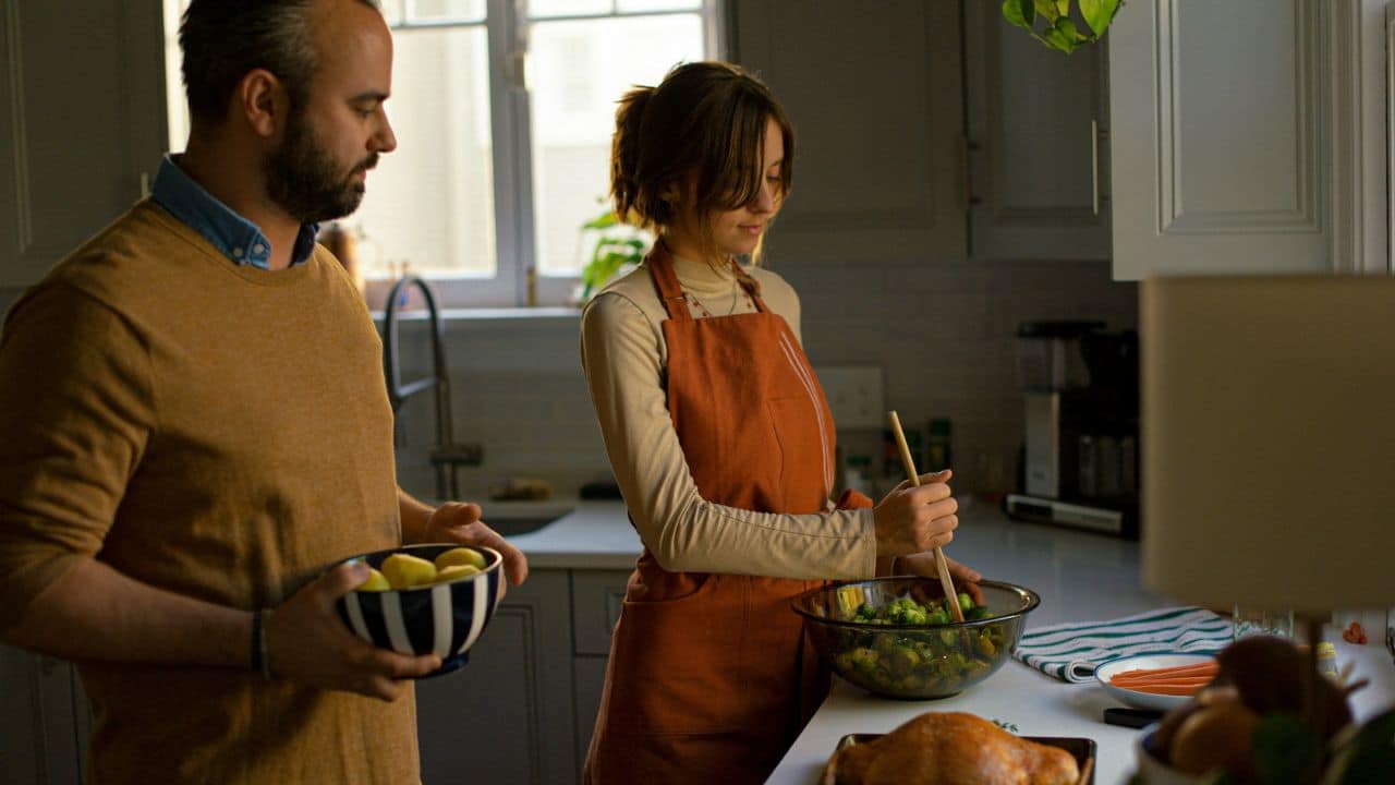 A couple cooking together in a kitchen, with the woman stirring a bowl and the man holding another bowl.